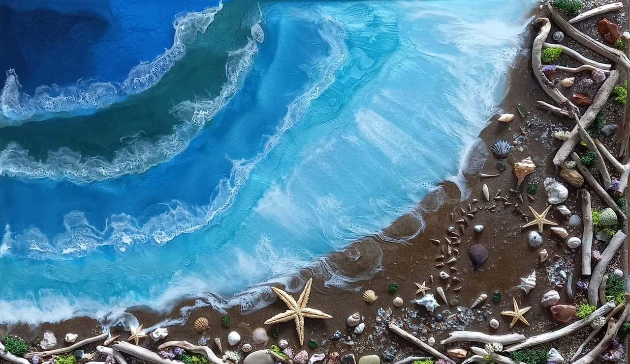 Resin-Bild von oben: Wellen mit weißer Gischt laufen auf dunklen Sand mit Seesternen, Muscheln und Treibholz